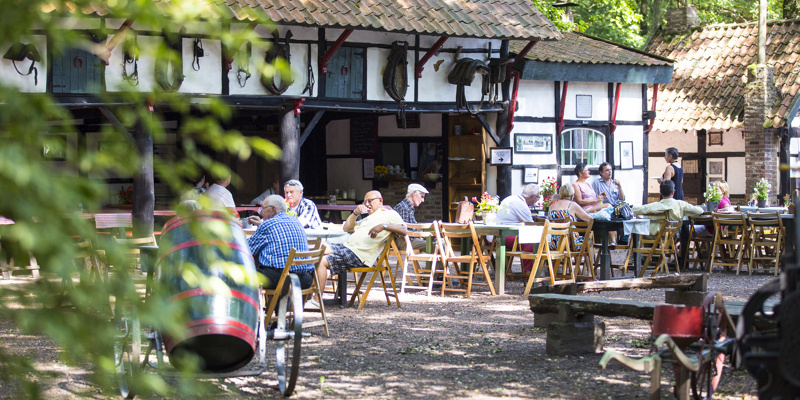 Mensen zitten op houten stoeltjes op het terras bij een oude vakwerkboerderij